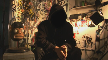 Szenenbild: Mann mit Kapuze auf, so dass sein Gesicht nicht zu sehen ist, vor Graffitti Wand