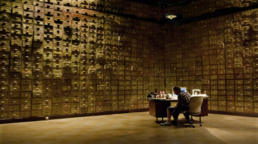 Szenenbild: Gott in seinem Arbeitszimmer, dessen Wände mit Schubladen übersäht sind