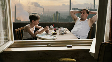 Szenenbild: Vater und Sohn am Frühstückstisch auf dem Balkon, im Hintergrund rauchende Schlote