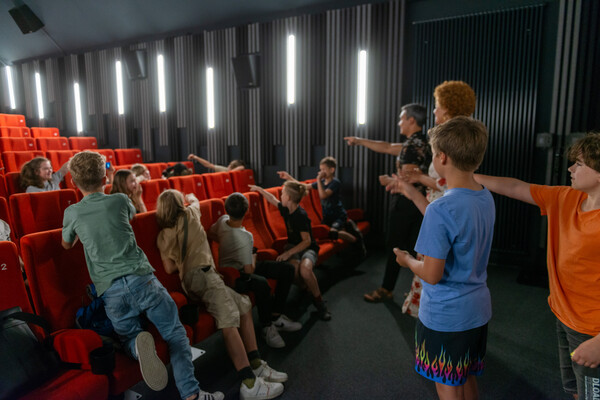 Im Kinosaal stehen zwei Kinder und zwei Erwachsene vor der Leinwand und zeigen nach hinten in den Saal, die anderen Kinder drehen sich auch nach hinten um