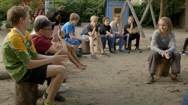 Szenenbild: Eine Gruppe von Kindern und Erwachsenen im Halbkreis auf einem Spielplatz sitzend