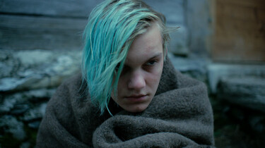 Szenenbild: Matteo, mit blau gefärbten Haaren, sitzt in eine graue Decke gehüllt vor der Hütte