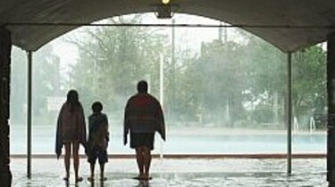 Szenenbild: Die Familie von hinten gesehen, sie schauen auf den im Regen liegenden Swimming-Pool