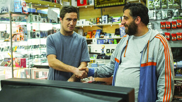 Szenenbild: Der Besitzer eines Ladens für gebrauchte Elektronik gibt sich vor einem Fernseher die Hand mit einem anderen Mann