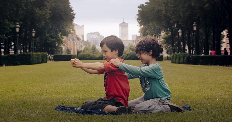 Filmstill "Die Götter von Molenbeek", Aatos und Amine sitzen hintereinander im Park auf der Wiese und gucken nach links, Arme auf Schulterhöhe gehoben, dahinter eine Stadtansicht, gerahmt von Bäumen 