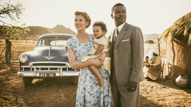 Szenenbild: Seretse und Ruth, mit Baby auf dem Arm in einem afrikanischen Dorf, im Hintergrund eine schwarze Limousine